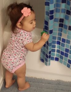 A baby girl in pink and white is playing with the shower curtain.